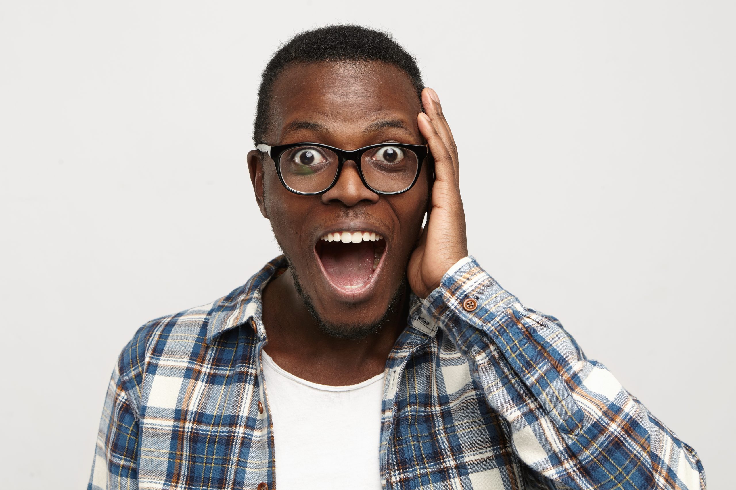 Amazed young African American hipster wearing glasses and checkered shirt over white t-shirt touching head and opening mouth widely, screaming with shock and disbelief, happy with promotion at work